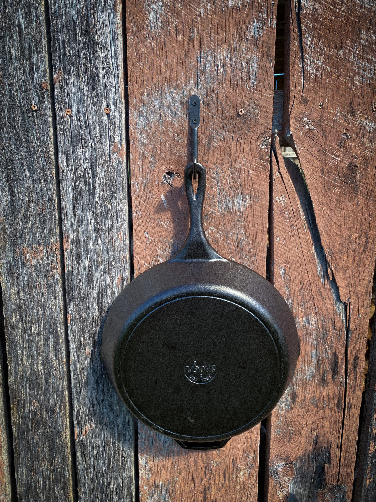 Hand-forged steel skillet hook holding a Lodge cast iron pan by the helper handle on a weathered wood wall. Hutton Forge.
