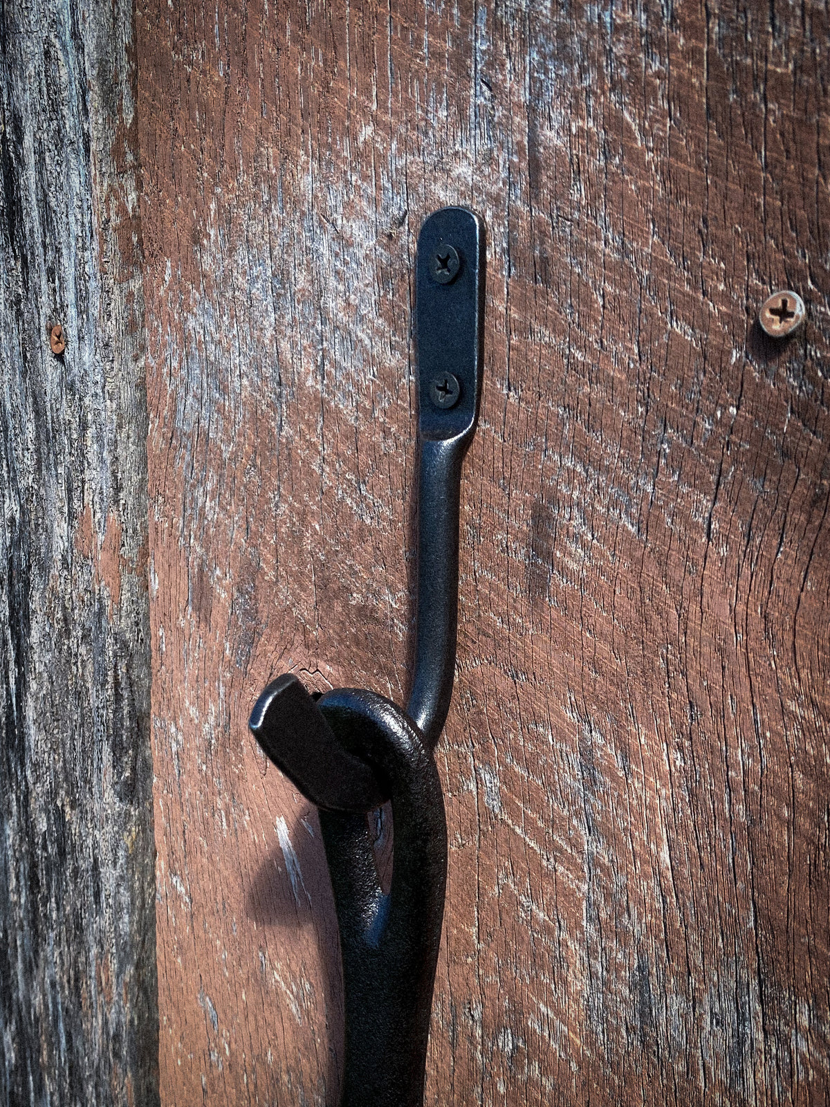 Hand-forged steel skillet hook holding a Lodge cast iron pan by the helper handle on a weathered wood wall. Hutton Forge.
