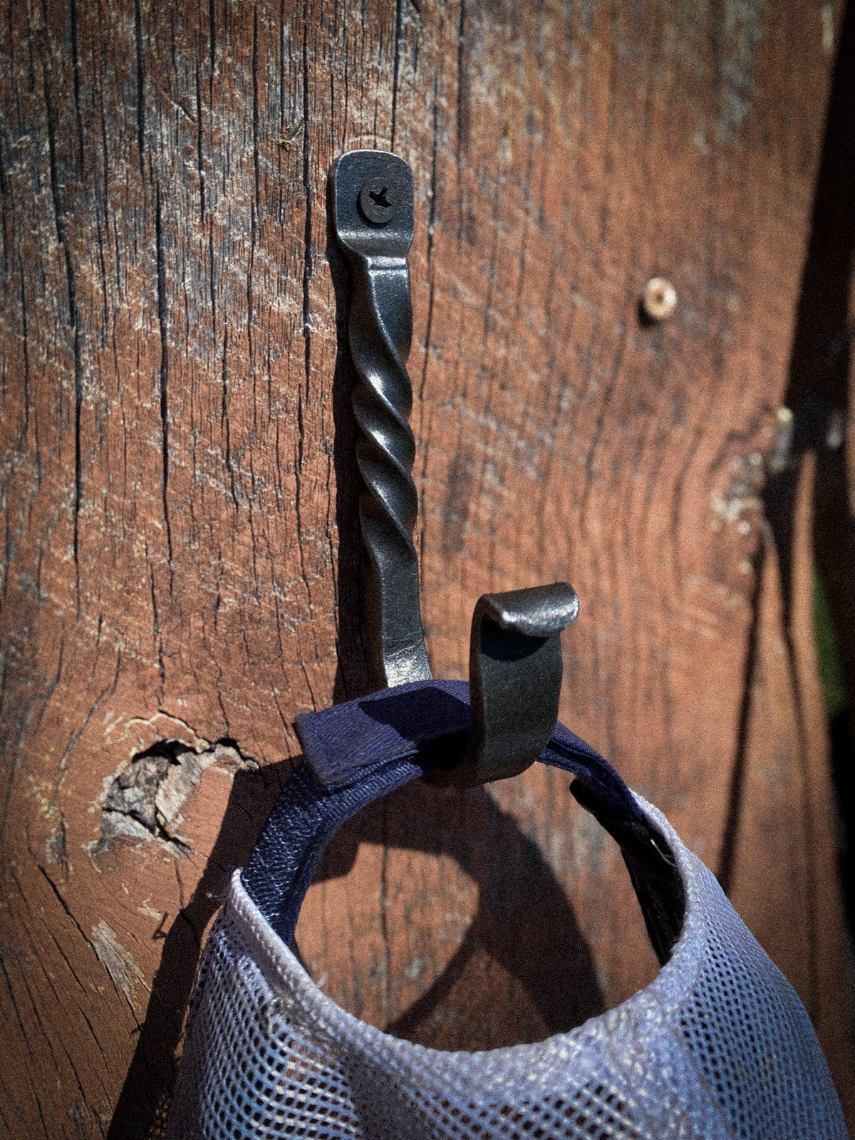 A black twisted metal wall hook hanging on a weathered brown wooden surface, holding a white baseball cap.