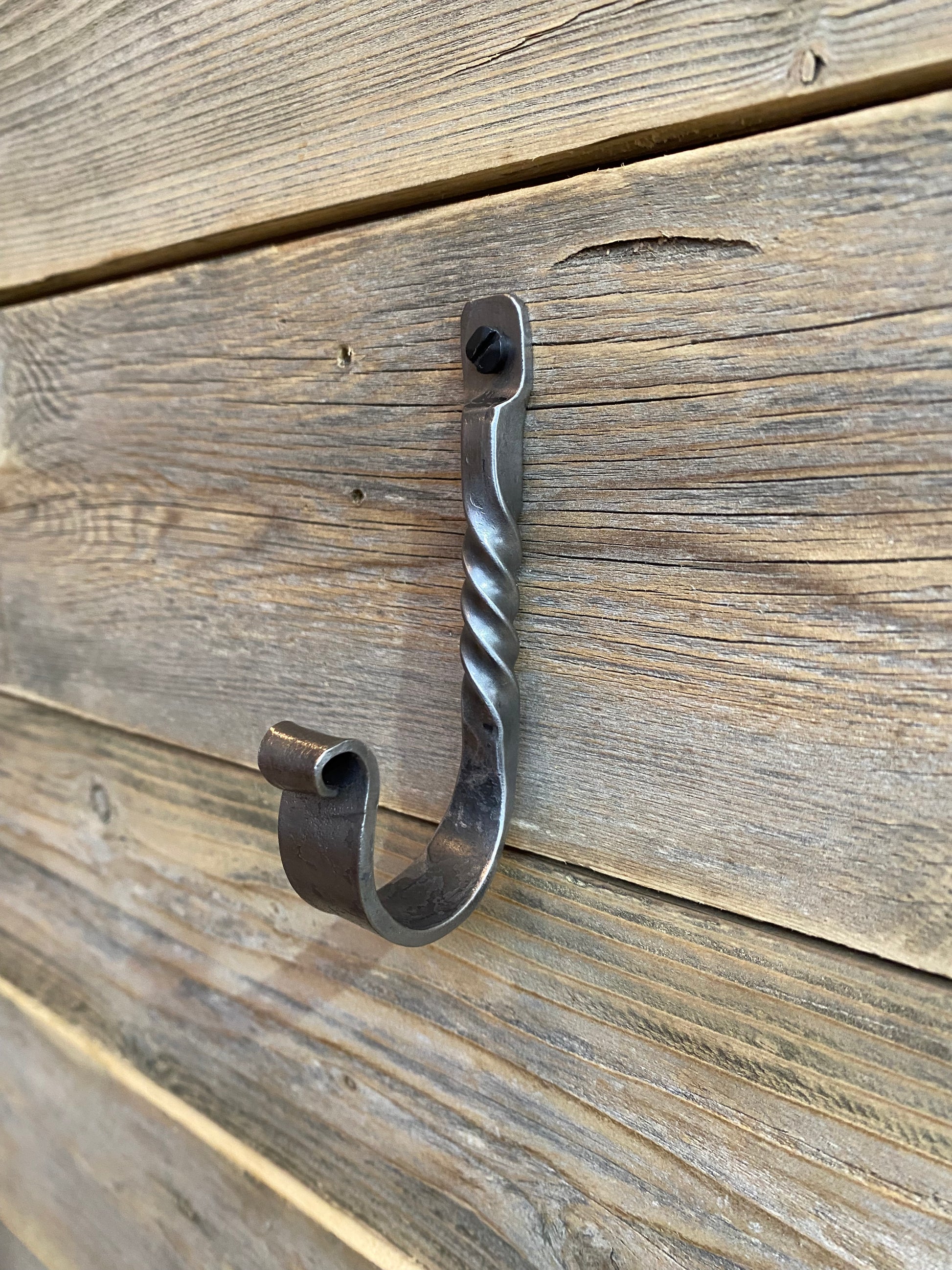 Hand-forged twisted steel J-hook in blackened finish, blacksmith-made with a decorative twist, shown on a rustic wooden surface. Hutton Forge.

