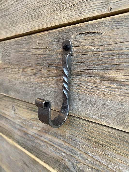 Hand-forged twisted steel J-hook in blackened finish, blacksmith-made with a decorative twist, shown on a rustic wooden surface. Hutton Forge.

