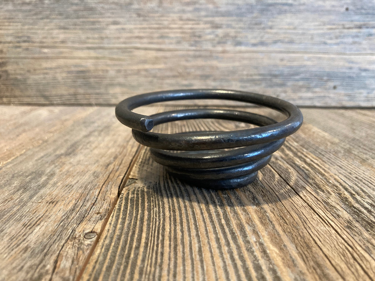 A dark metallic coiled candle holder, resembling a small spring or decorative sculpture, resting on a rustic wooden surface with a matching wood plank background.