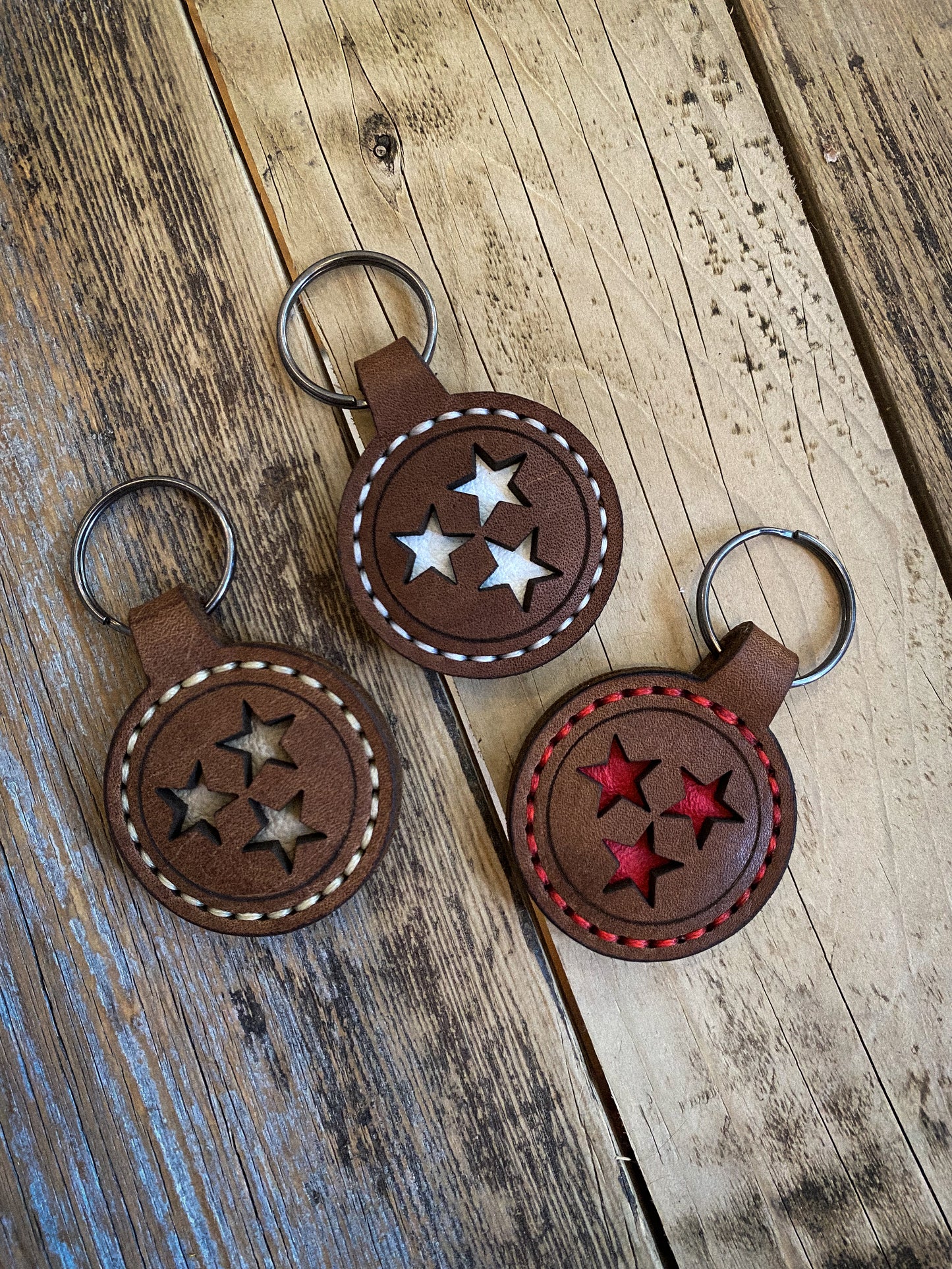 Three hand-stitched brown leather keychains featuring the Tennessee Tri-Star design with cut-out stars, shown in different stitching colors on a rustic wooden surface. Hutton Forge.

