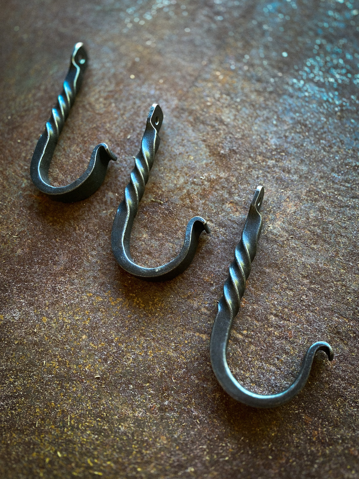 Three rustic black metal J-hooks with twisted spiral details, arranged diagonally on a textured, rusty brown surface.