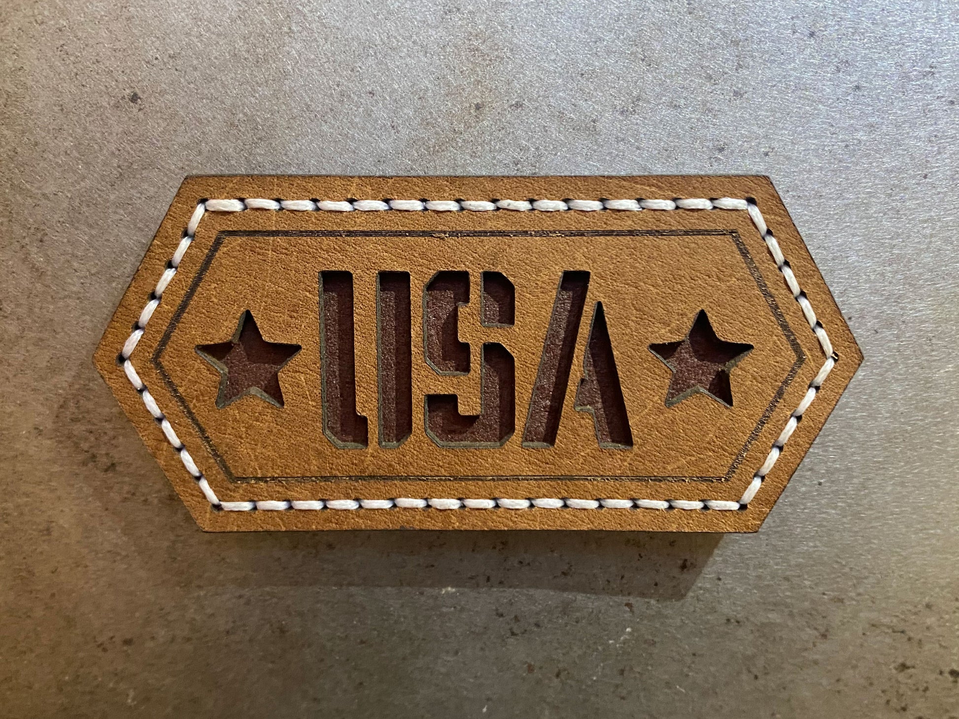 Hand-stitched brown leather magnet with cut-out “USA” lettering and stars, featuring white stitching on a hexagonal design. Hutton Forge.
