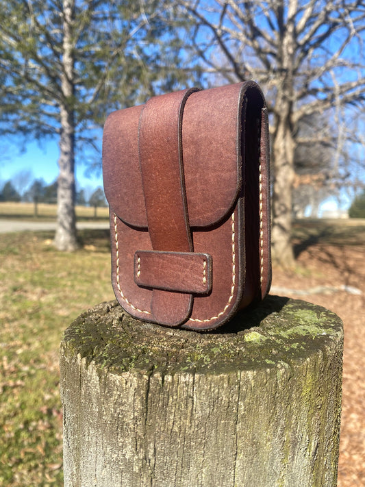 Handmade brown leather pouch with fold-over flap, vertical strap, and hand stitching, standing on a weathered wooden post outdoors under a clear blue sky. Hutton Forge belt pouch.

