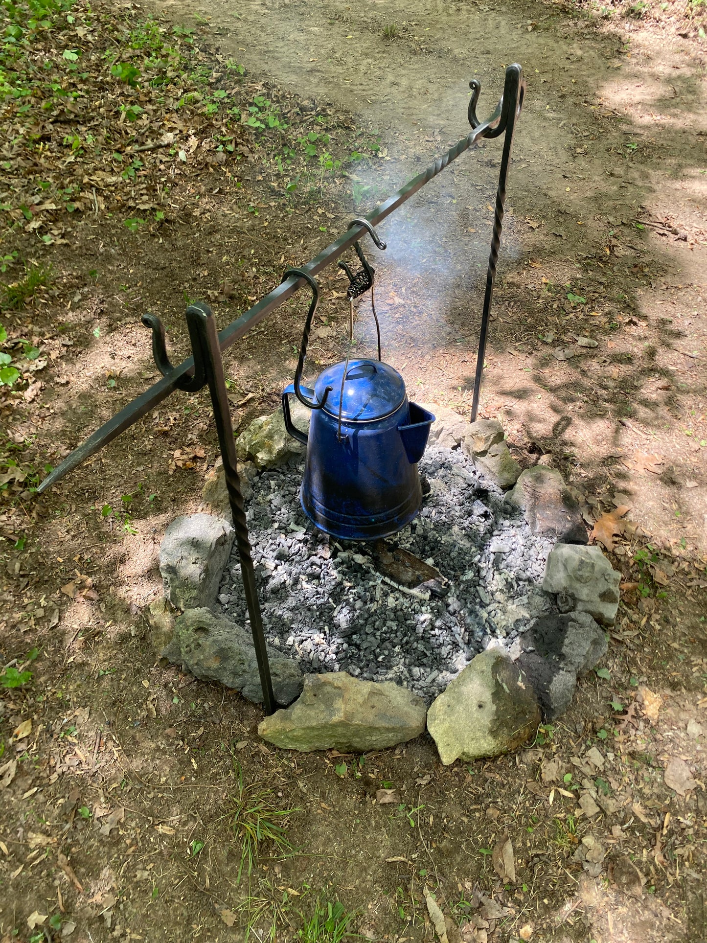 A blue metal kettle or pot hangs from a black metal tripod stand over an outdoor stone fire pit with ash and embers, set on dirt ground.