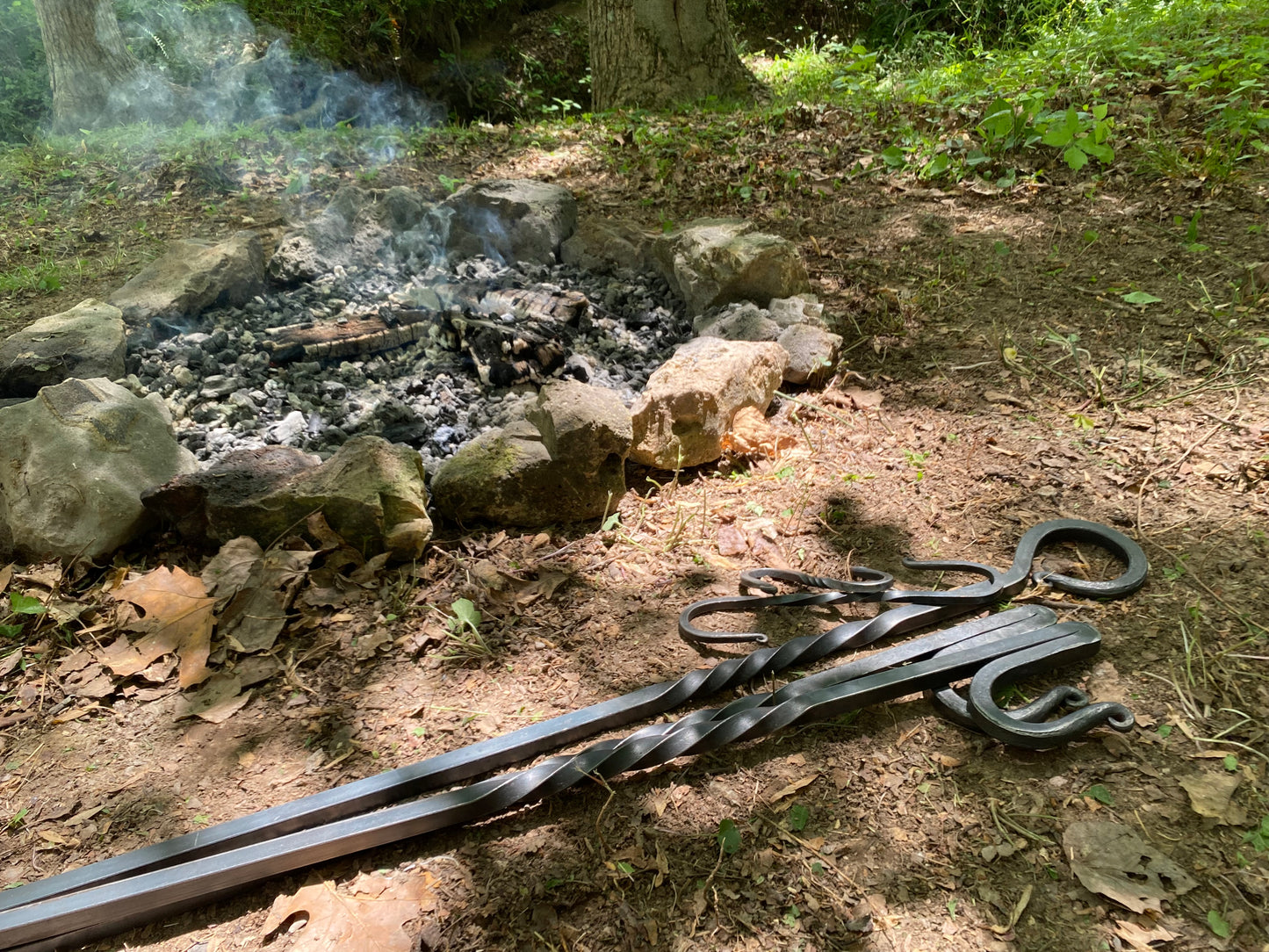 The dismantled tripod, resting on the ground in a forest clearing with a stone fire pit and embers in the background.