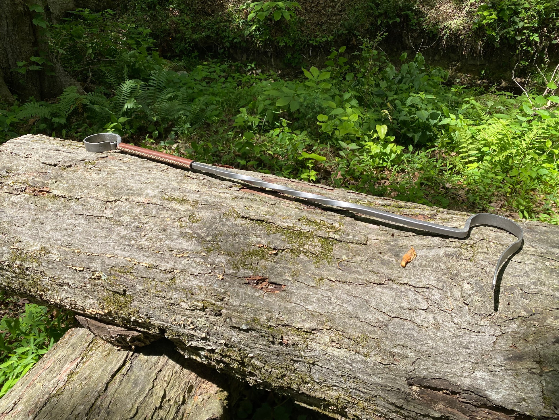 A long-handled steel fire poker, resting on a large weathered log in a forest setting with green foliage in the background.