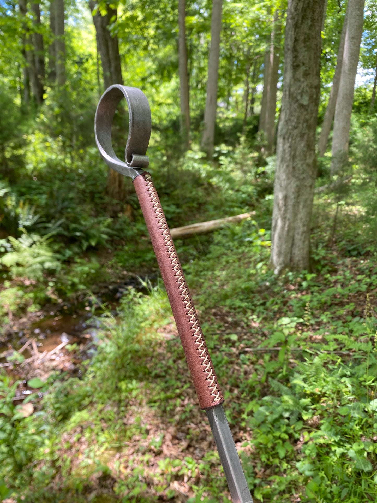 A close-up of a handcrafted steel fire poker with a curved loop end and a brown leather-wrapped handle, set against a blurred forest background.