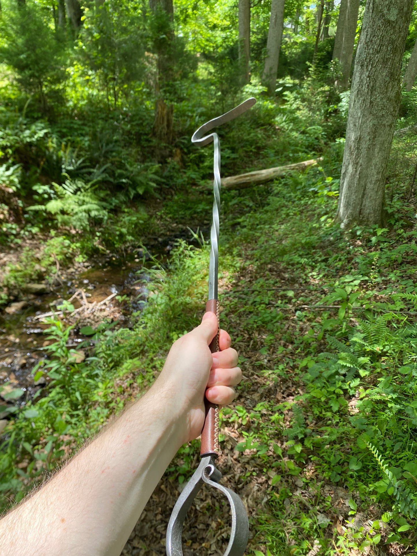 Close-up of a leather-wrapped fire poker with a hand-forged steel ring end and brown leather handle with visible stitching, lying on a weathered log surrounded by green ferns. Hutton Forge.

