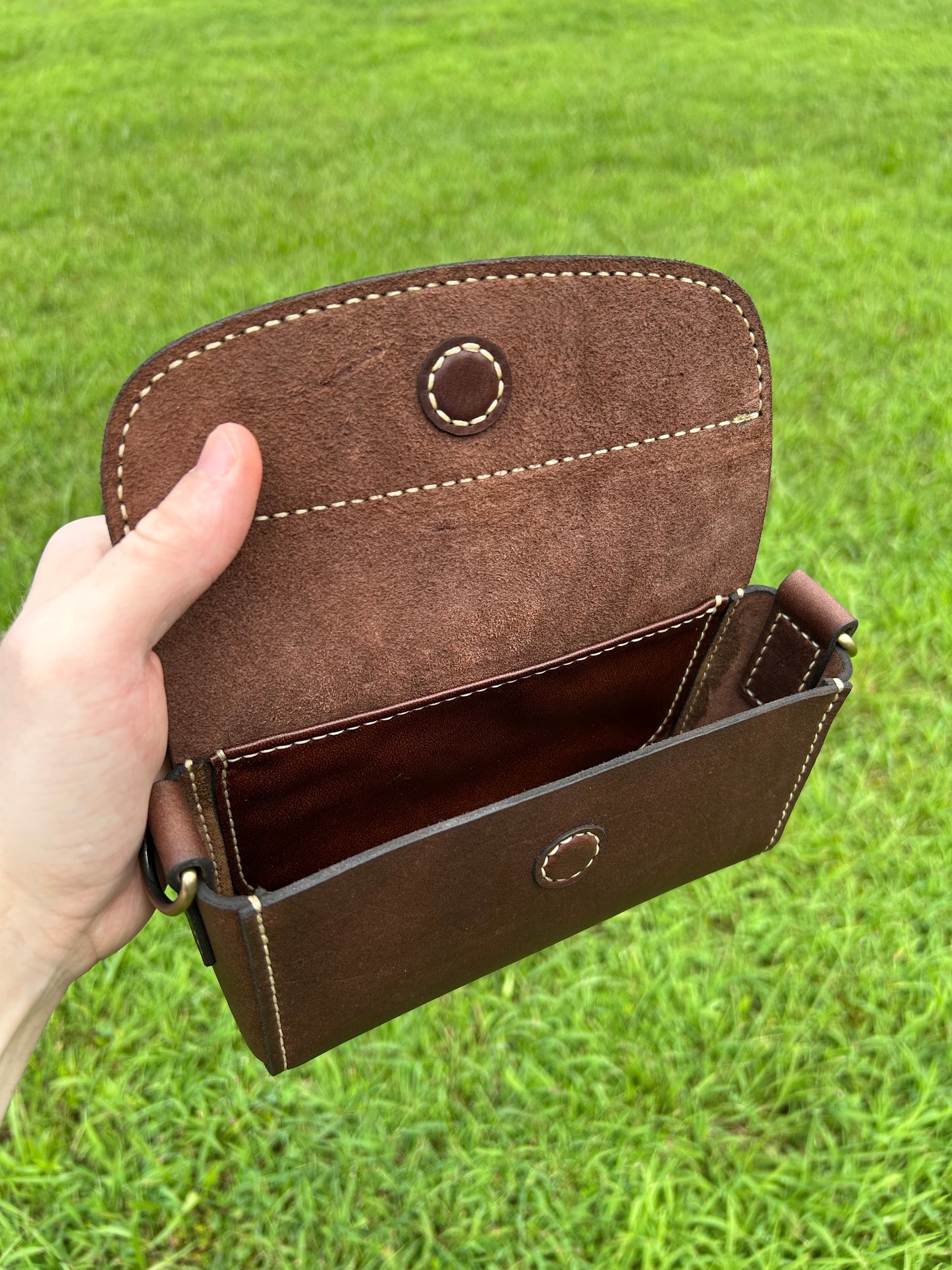 A brown leather purse held open by a hand against a green grass background. The purse has a flap closure with a magnetic button, contrast stitching, and a visible interior compartment. Made by the leathersmith at Hutton Forge. 