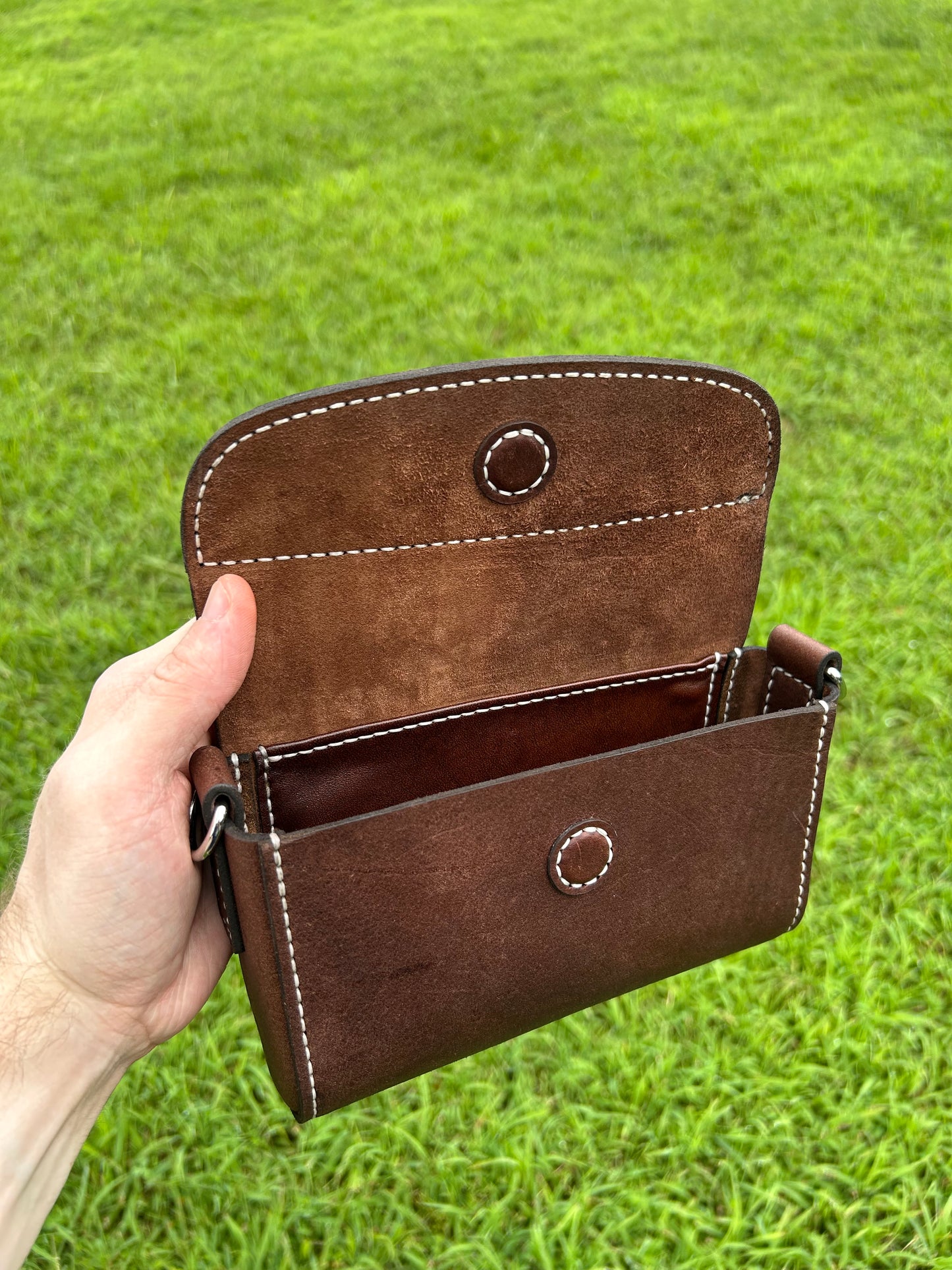 A hand holding an open brown leather purse  against a green grass background. The pouch has visible white contrast stitching, circular metal snap details, and a dark interior.