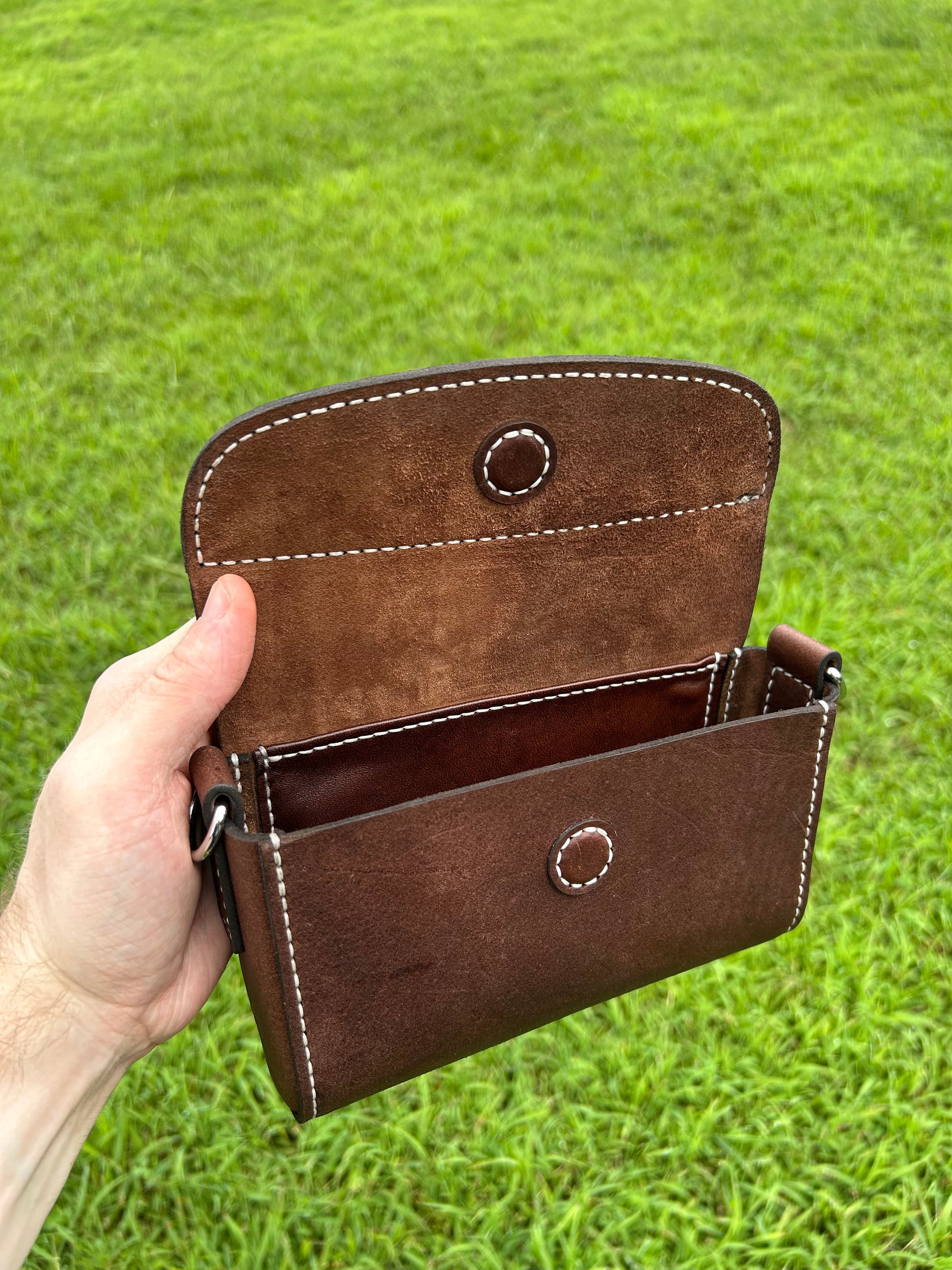 A hand holding an open brown leather purse  against a green grass background. The pouch has visible white contrast stitching, circular metal snap details, and a dark interior.