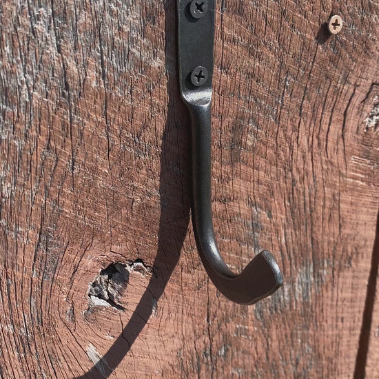 Hand-forged steel skillet hook holding a Lodge cast iron pan by the helper handle on a weathered wood wall. Hutton Forge.
