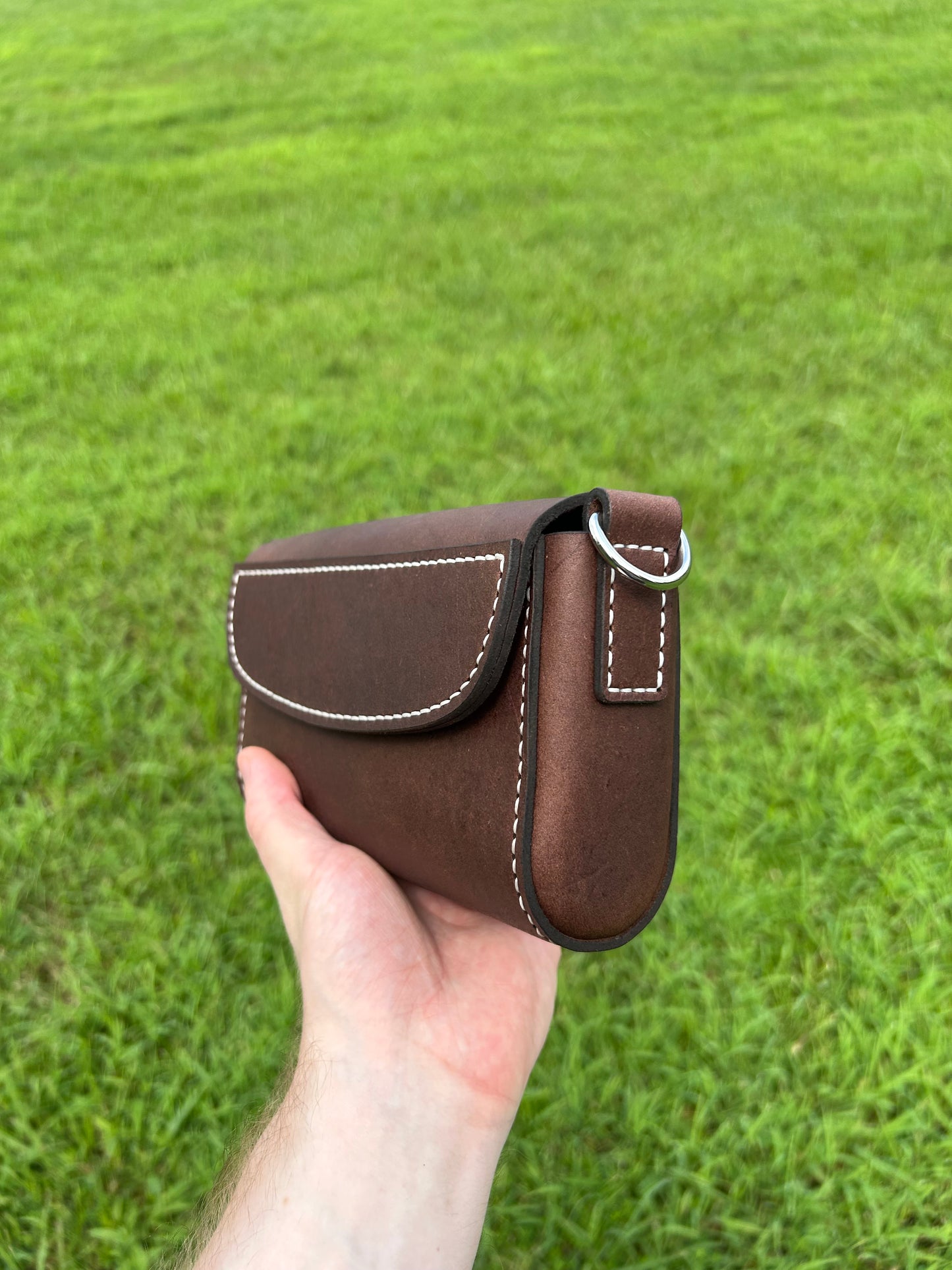A hand holding a small brown leather purse with white contrast stitching and a silver metal ring, set against a blurred green grass background.