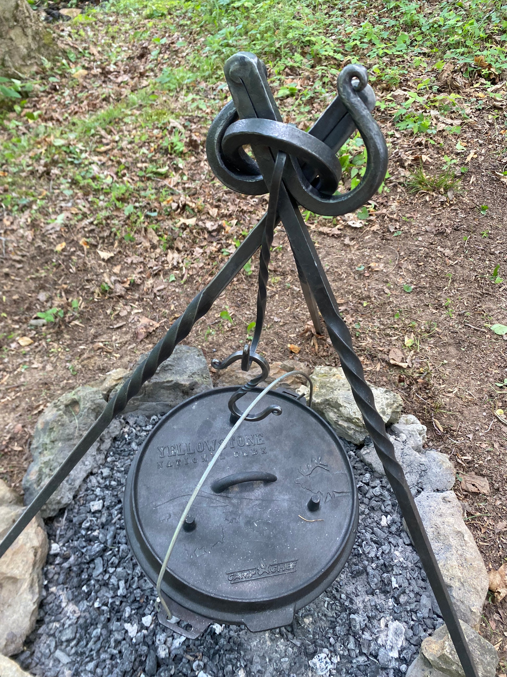 A black cast iron dutch oven with a twisted handle and hanging hook, suspended from a tripod stand outdoors over a stone fire pit.