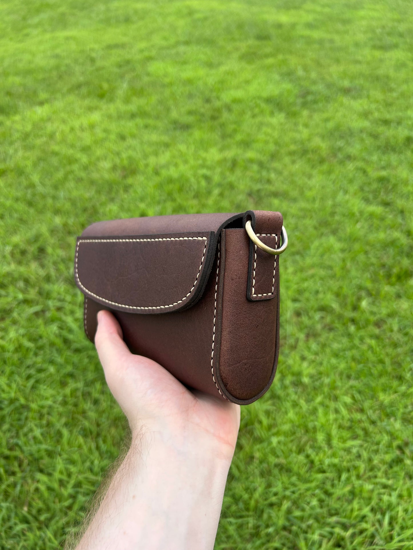 Hand holding a small brown leather crossbody purse with flap closure and hand stitching. Made by the leathersmith at Hutton Forge.
