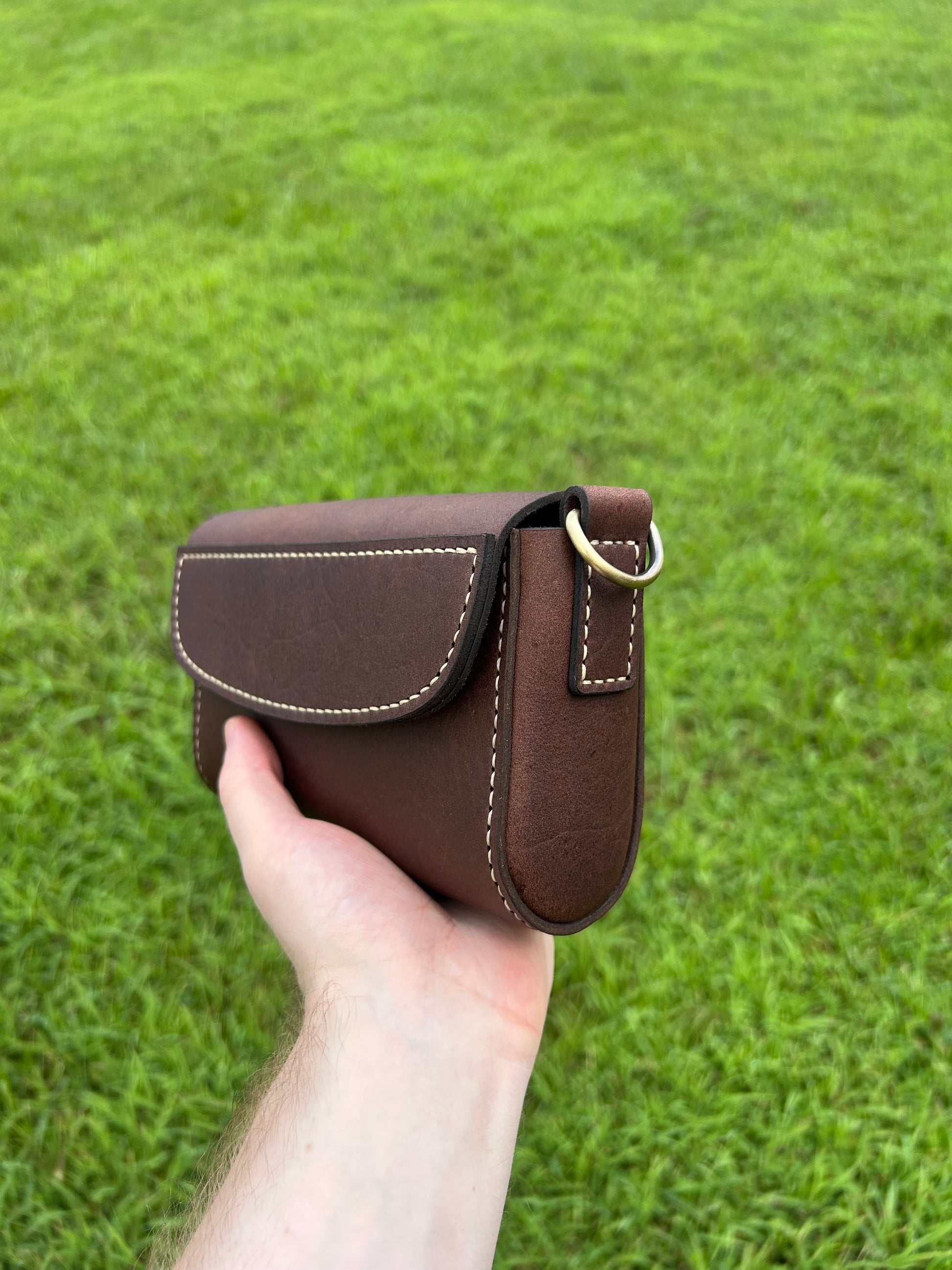 Hand holding a small brown leather crossbody purse with flap closure and hand stitching. Made by the leathersmith at Hutton Forge.
